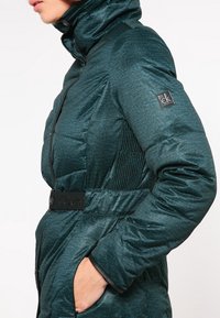 Tyrkysová péřová bunda s vysokým límcem, s texturovaným povrchem, elastickými bočními panely a černým páskem s logem.