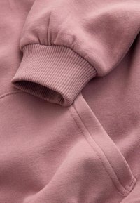 Sudadera rosa con puños acanalados. La tela es suave y texturizada, con un brillo sutil. Entre los detalles destacados se encuentran las costuras y el elástico en los puños.