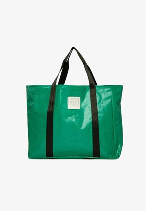 Zaļš iepirkuma soma, izgatavota no teksturēta, izturīga materiāla ar melnām rokām. Priekšpusē ir kvadrātveida balta logotipa plāksne.