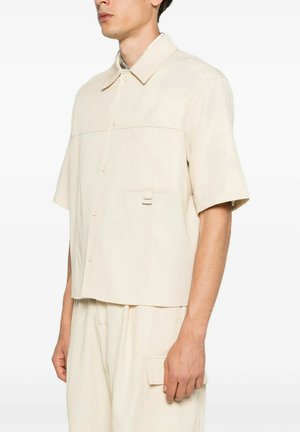 Uomo che indossa una camicia beige, ampia e a maniche corte, con bottoni e tasca frontale, e pantaloni beige coordinati con tasca laterale.