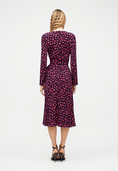 Γυναίκα με κυματιστά ξανθά μαλλιά που φοράει μεσαίο φορεμα σε ναυτικό μπλε με κόκκινο λουλουδένιο σχέδιο και μαύρα ψηλοτάκουνα σανδάλια, γυρισμένη προς τα πίσω.