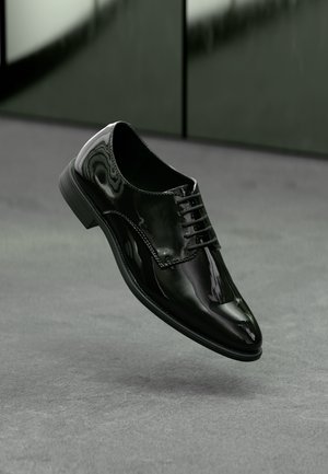 Chaussure habillée en cuir noir brillant avec lacets, flottant au-dessus d’une surface grise avec un arrière-plan sombre et flou.