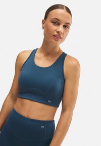 Top corto atlético en verde azulado oscuro, con un escote redondeado, detalles de costura y el logotipo de la marca en el dobladillo. Material suave y elástico.