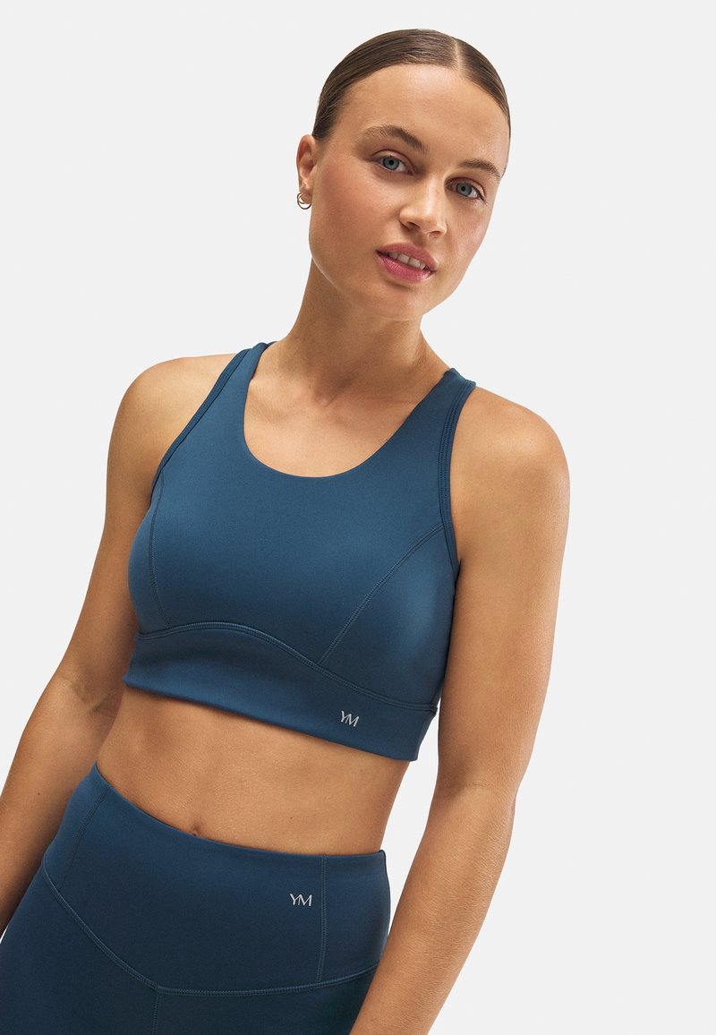 Top corto atlético en verde azulado oscuro, con un escote redondeado, detalles de costura y el logotipo de la marca en el dobladillo. Material suave y elástico.