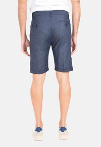 Pantaloni corti blu navy, lunghi fino al ginocchio, realizzati in tessuto testurizzato. Presentano due tasche posteriori con bottoni e orli arrotolati, indossati con scarpe da ginnastica chiare.