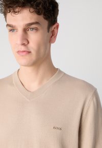Beige V-nyakú pulóver bordázott gallérral, puha anyagból készült, diszkrét hímzett logóval a mellrészen.
