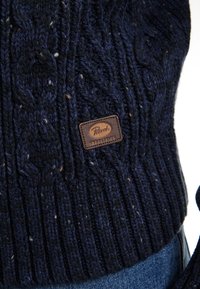 Pulover din tricot cu model împletit, de culoare bleumarin, cu tiv zimțat. Are un eticheta din piele în partea de jos, cu inscripția "Petrol Industries" debossată. Texturat și călduros.