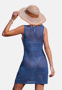 Robe en crochet bleu foncé, sans manches avec des motifs ajourés. Associée à un chapeau en paille à large bord et des bracelets.