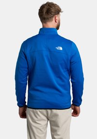 Veste en polaire bleue avec un col montant, des manches longues et une bordure noire au bas. Logo au dos. Texture lisse et couleur unie.