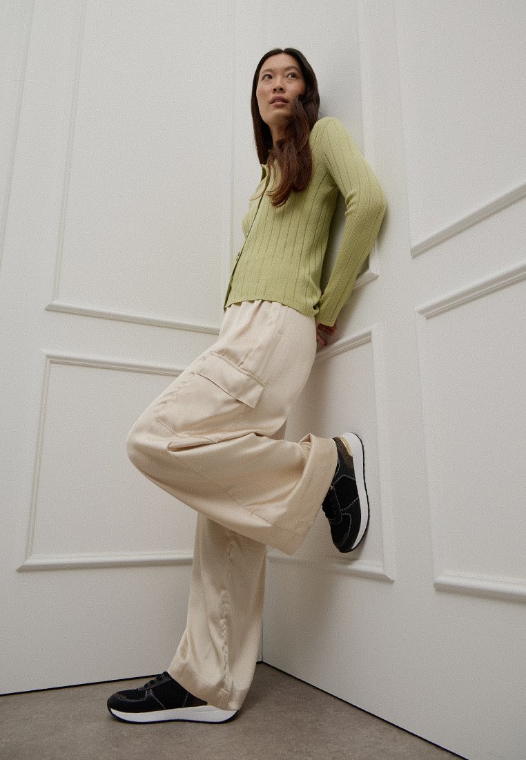 Una mujer joven con un suéter de canalé verde claro y pantalones cargo beige se apoya contra una pared blanca panelada, vistiendo zapatillas negras.