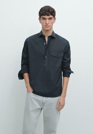 Junger Mann mit kurzen lockigen Haaren, der ein schwarzes Langarmhemd mit Brusttasche und hellgraue Hose trägt, die Hände in den Taschen.