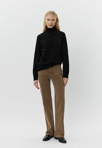 Maglione nero a collo alto, maniche lunghe. Jeans marroni a campana con tasche frontali e dettagli ricamati. Scarpe nere. Sfondo neutro.