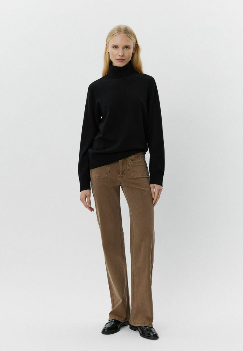 Maglione nero a collo alto, maniche lunghe. Jeans marroni a campana con tasche frontali e dettagli ricamati. Scarpe nere. Sfondo neutro.