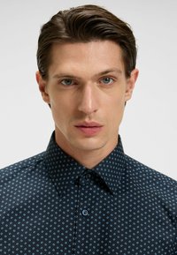 Tummansininen napapaita, jossa on vaaleansininen geometrinen kuvio. Ominaisuuksiin kuuluu levitetty kaulus ja kontrastiväriset siniset napit. Sileä pinta.