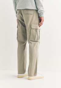 Pantalon cargo beige avec poches latérales, détails zippés et coupe décontractée. Porté avec des baskets blanches, mettant en valeur une texture en coton lisse.