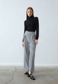 Črn prilegajoč pulover z visokim ovratnikom, široke hlače v houndstooth vzorcu s sprednjimi gubami in dekorativnimi zlatimi gumbki, v kombinaciji s črnimi loafers z blok peto.