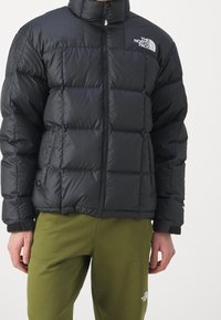 Man i en svart vadderad jacka från The North Face med dragkedjan uppe och olivgröna byxor från The North Face mot en enfärgad bakgrund.