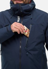 Navy blue waterproof jacket with high collar, zippered chest pocket, and adjustable drawstrings; featuring logo on the front and textured material.