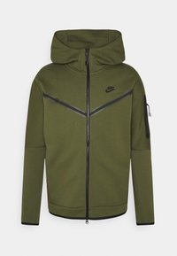 Olivgrön zip-up hoodie med dragsko i huvan, svarta accenter och diagonala ränder på bröstet. Har sidoficka med dragkedja och mjuk textur.