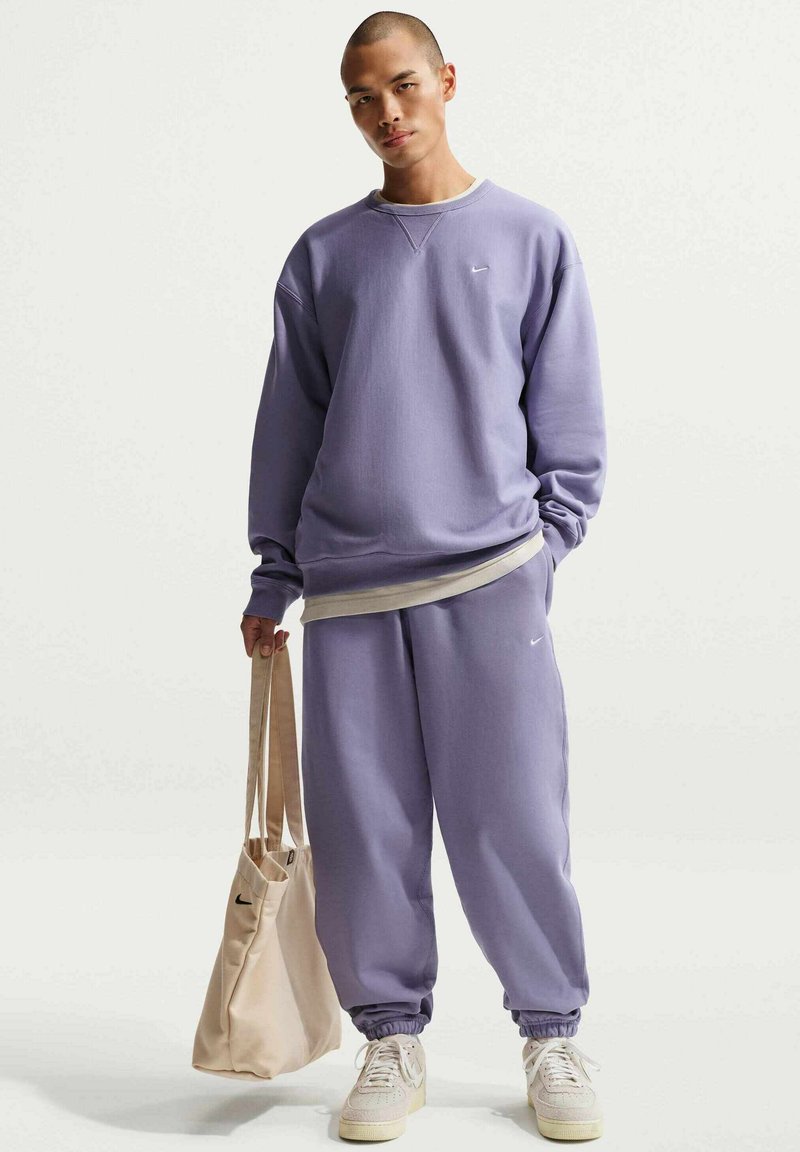 Sweat-shirt violet et jogging assorti en tissu doux. Comprend des poignets élastiques, un col rond et un petit logo. Sac fourre-tout beige porté sur une épaule.