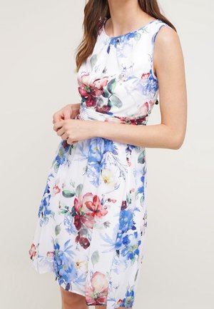 Woman wearing a sleeveless white dress with red, blue, and green floral patterns, standing with hands clasped at waist.