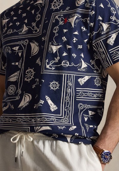 Camicia blu navy con motivi nautici bianchi tra cui barche a vela, ancore, fari, corde e timoni, indossata con pantaloni bianchi con coulisse.