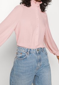 Blusa rosa chiaro a maniche lunghe con colletto arricciato e chiusura con bottoni, abbinata a jeans azzurri a vita alta. Tessuto morbido.