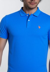 En man som bär en figursydd, kortärmad ljusblå poloach med en liten orange logotyp på vänster bröst. Bakgrunden är neutral.