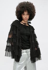 Sort tylkimono med lagdelte flæser, der har bindelukning ved halsen. Kombineret med en sort top og sølvfarvede metallicbukser.