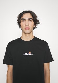 Sort bomulds T-shirt med et lille hvidt og rødt broderet logo på brystet, klassisk rund hals og korte ærmer.