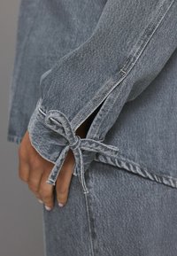 Hellgraue Jeansjacke mit einem Bindeband am Ärmelbund, versehen mit fransigen Kanten und kontrastierenden Nähten entlang der Nähte.