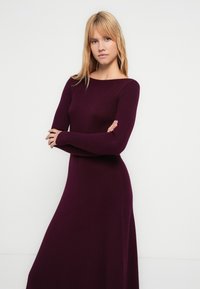 Lange paarse geribbelde jurk met een boothals en lange mouwen, met een getailleerd bovenstuk en een uitlopende onderkant. Gladde textuur, minimale details.