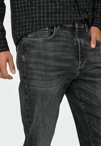 Tamsiai pilkos spalvos džinsai iš džinsinio audinio, turintys klasikinį penkių kišenių dizainą ir šiek tiek išblukusį atspalvį, derinami su juoda ir žalia languota marškinėliais.