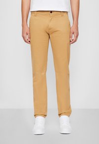 Pantalons en coton beige avec un design à jambes droites, featuring des poches avant, une fermeture à bouton, et portés avec des baskets blanches. Texture lisse.