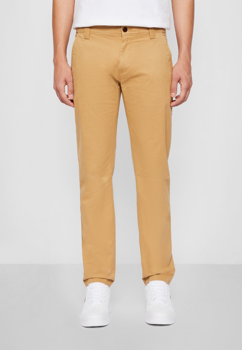 Pantalons en coton beige avec un design à jambes droites, featuring des poches avant, une fermeture à bouton, et portés avec des baskets blanches. Texture lisse.
