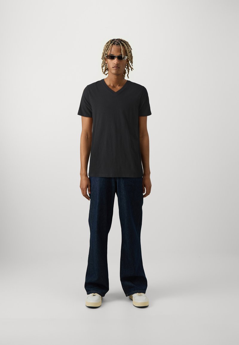 Schwarzes Baumwoll-V-Ausschnitt-T-Shirt, lockere dunkle Jeanshose und weiße Sneaker. Das Model trägt dunkle Sonnenbrillen und hat schulterlanges, strukturiertes Haar.