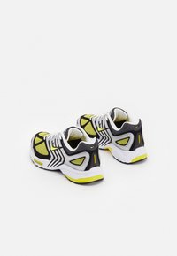 Svartvita sportskor med gula accenter, nätpaneler, vågmönster och en dämpad sula. Har en rund tå och snörningsdesign.