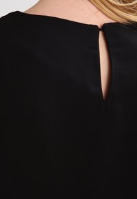 Arrière du cou d'une personne aux cheveux blonds, portant un vêtement noir avec une petite ouverture en forme de trou de serrure au niveau du col.