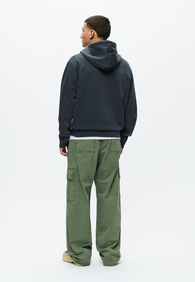 Mann von hinten, trägt einen dunklen Kapuzenpullover, grüne Cargo-Hosen und beige Schuhe vor einem schlichten hellen Hintergrund.