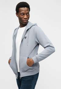 Άνδρας με ανοιχτό μπλε φούτερ με φερμουάρ και τσέπες, λευκό μπλουζάκι και σκούρο τζιν, που κοιτάει πλάγια σε απλό φόντο.