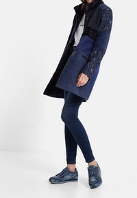Cappotto blu strutturato con colletto nero, caratterizzato da chiusura con bottoni, abbinato a jeans denim scuri e sneakers blu a fantasia.