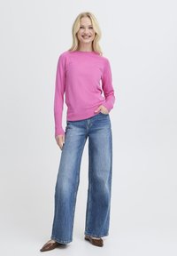 Femme blonde souriante portant un pull long à manches longues rose vif, un jean bleu à jambes larges et des chaussures pointues marron, se tenant devant un fond blanc.