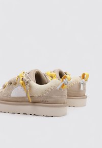 Par de zapatillas de ante beige con cordones amarillos y grises, suelas gruesas beige y detalles de costura, mostradas desde los ángulos trasero y lateral.