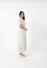 Abito maxi bianco con maniche corte a sbuffo e motivo floreale in rosa e verde. La modella indossa décolleté con open-toe, in piedi con le mani unite.