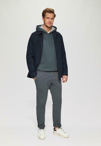 Veste marine sur un sweat à capuche vert, pantalon gris et baskets blanches. La tenue présente un design décontracté avec des textures douces et des manches retroussées.