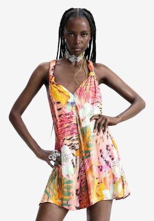 Donna con capelli intrecciati indossa un vestito estivo floreale colorato e una collana a forma di fiore d'argento, posando con le mani sui fianchi su uno sfondo bianco.