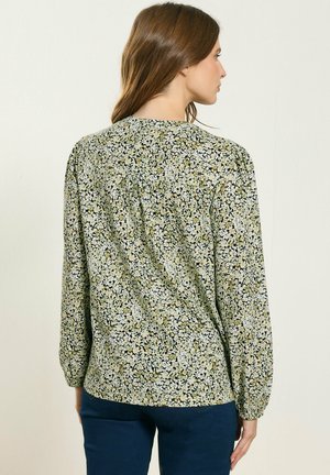 Nainen selin, yllään vihreä- ja valkokukkainen pitkähihainen paita ja tummansiniset housut, taustalla vaalea yksinkertainen seinä.