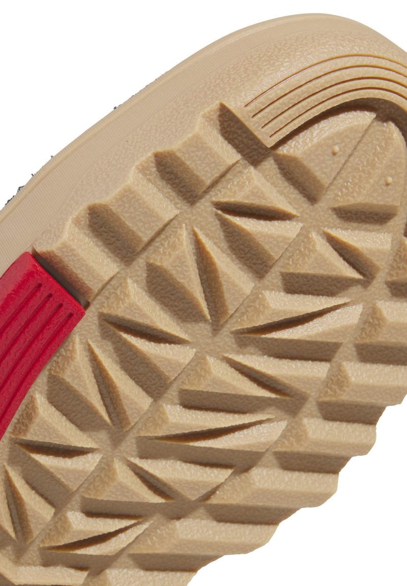 Sko)såler lavet af beige gummi, der har en struktureret overflade og trekantede mønstre, accentueret med en rød stribe.