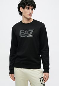 Fekete pamutkeverék anyagú pulóver, kerek nyakkivágással és hosszú ujjakkal. Elülső részén fehér grafikus logo található: "EA7 Emporio Armani."