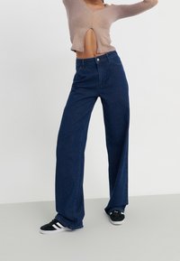 Persona que lleva jeans de pierna ancha de color azul oscuro, zapatillas negras y blancas, y un top cropped de manga larga beige con un corte frontal, de pie con los brazos levantados.